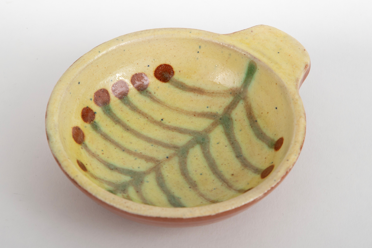 Kabaretskål av dreid leirgods. Skålene er runde med en enkel flat hank. På innsiden har skålen gulhvit glasur og er dekorert med en grønne gren.
