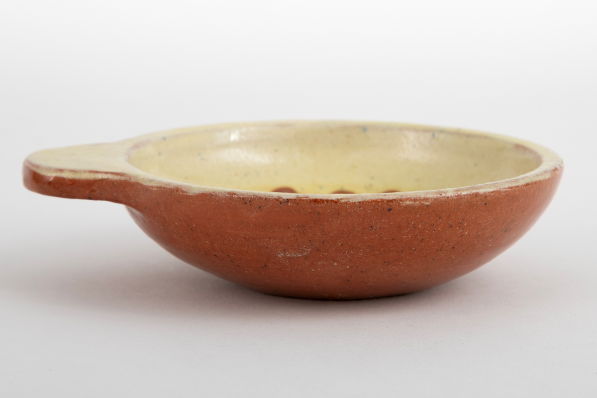 Kabaretskål av dreid leirgods. Skålene er runde med en enkel flat hank. På innsiden har skålen gulhvit glasur og er dekorert med en grønne gren.
