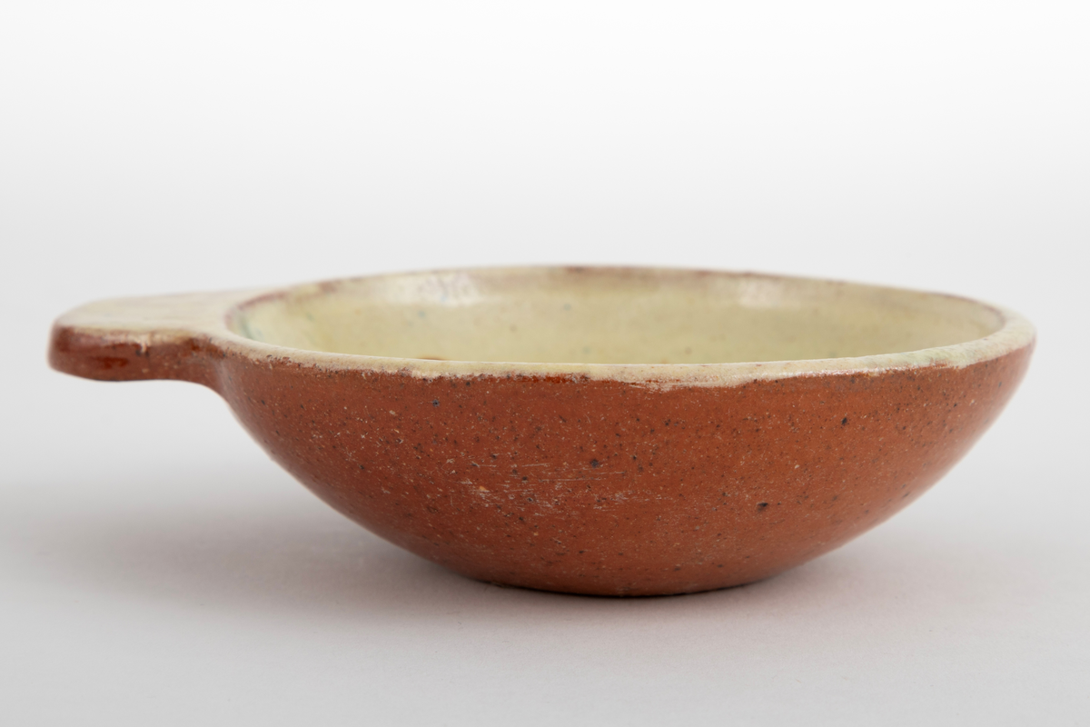 Kabaretskål av dreid leirgods. Skålene er runde med en enkel flat hank. På innsiden har skålen gulhvit glasur og er dekorert med en grønne gren.
