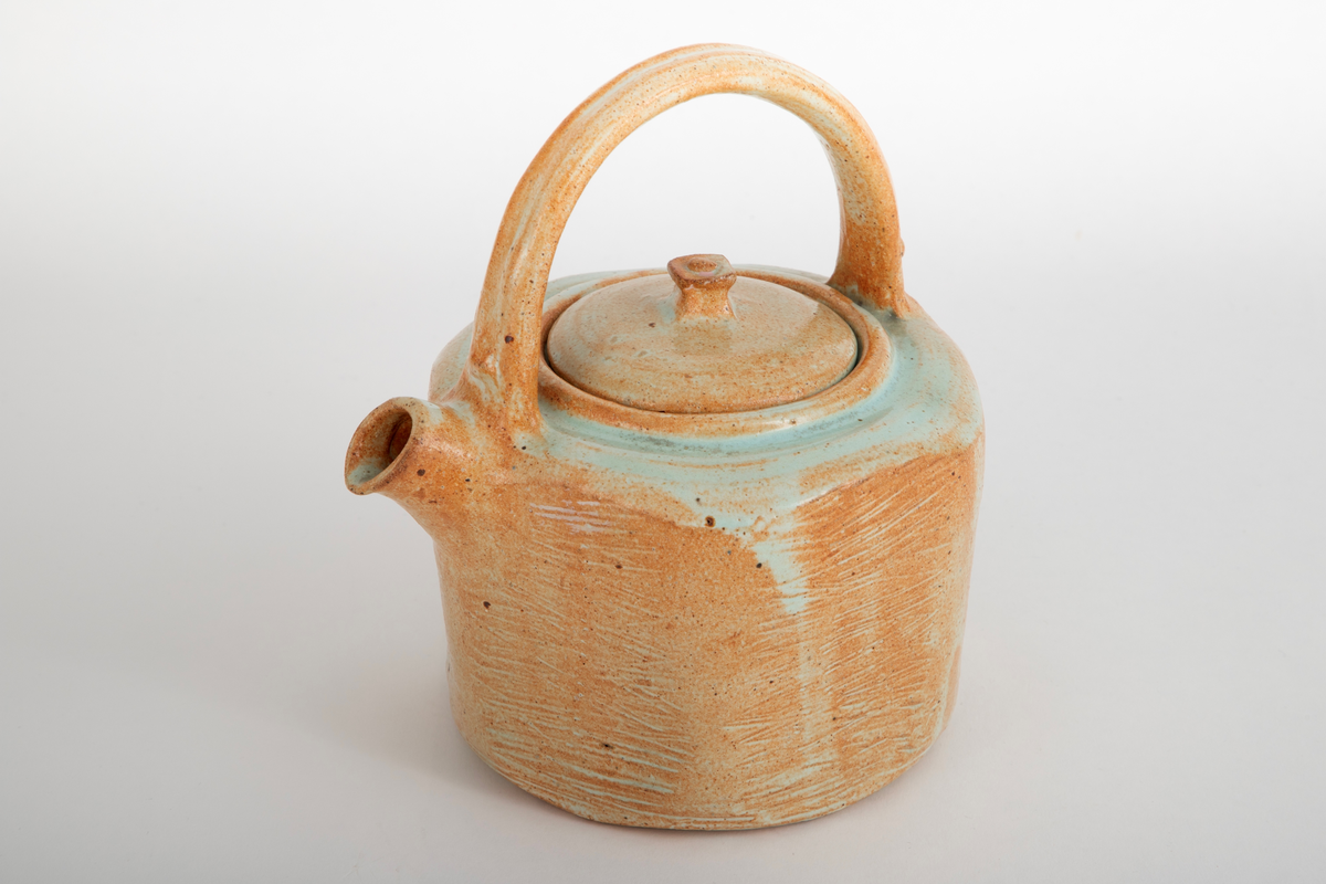 Åttekantet tekanne i steingods med begitning og glasur. Grønn glasur på toppen av kannen. Kannen er banket til for å få den den åttekantet formen. På sidene er det banket inn riller. Firkantet knopp på lokket.
