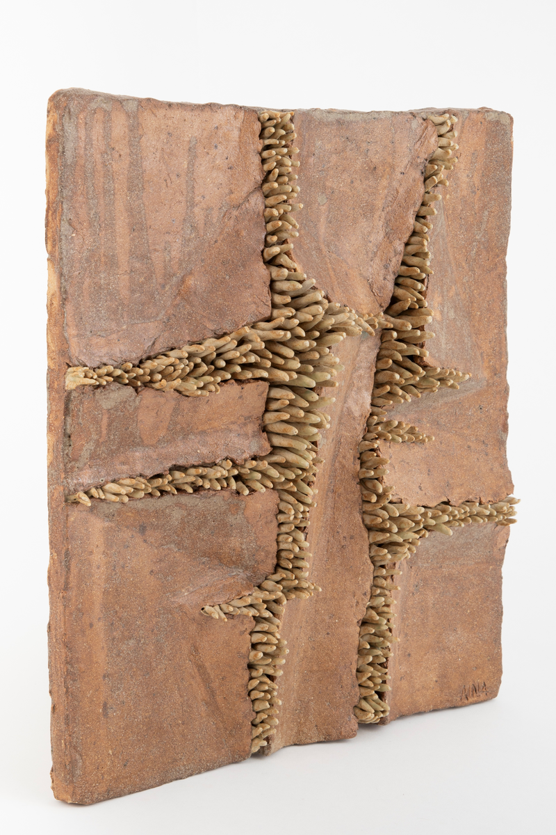 Kvadratisk plate i glasert steingods. På platen ser vi "tentakler" som bryter seg gjennom leira, hever den og danner sprekker i flaten. Utmeislet på undersiden med feste for oppheng.