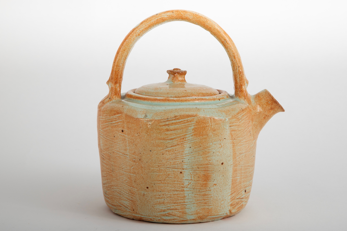 Åttekantet tekanne i steingods med begitning og glasur. Grønn glasur på toppen av kannen. Kannen er banket til for å få den den åttekantet formen. På sidene er det banket inn riller. Firkantet knopp på lokket.
