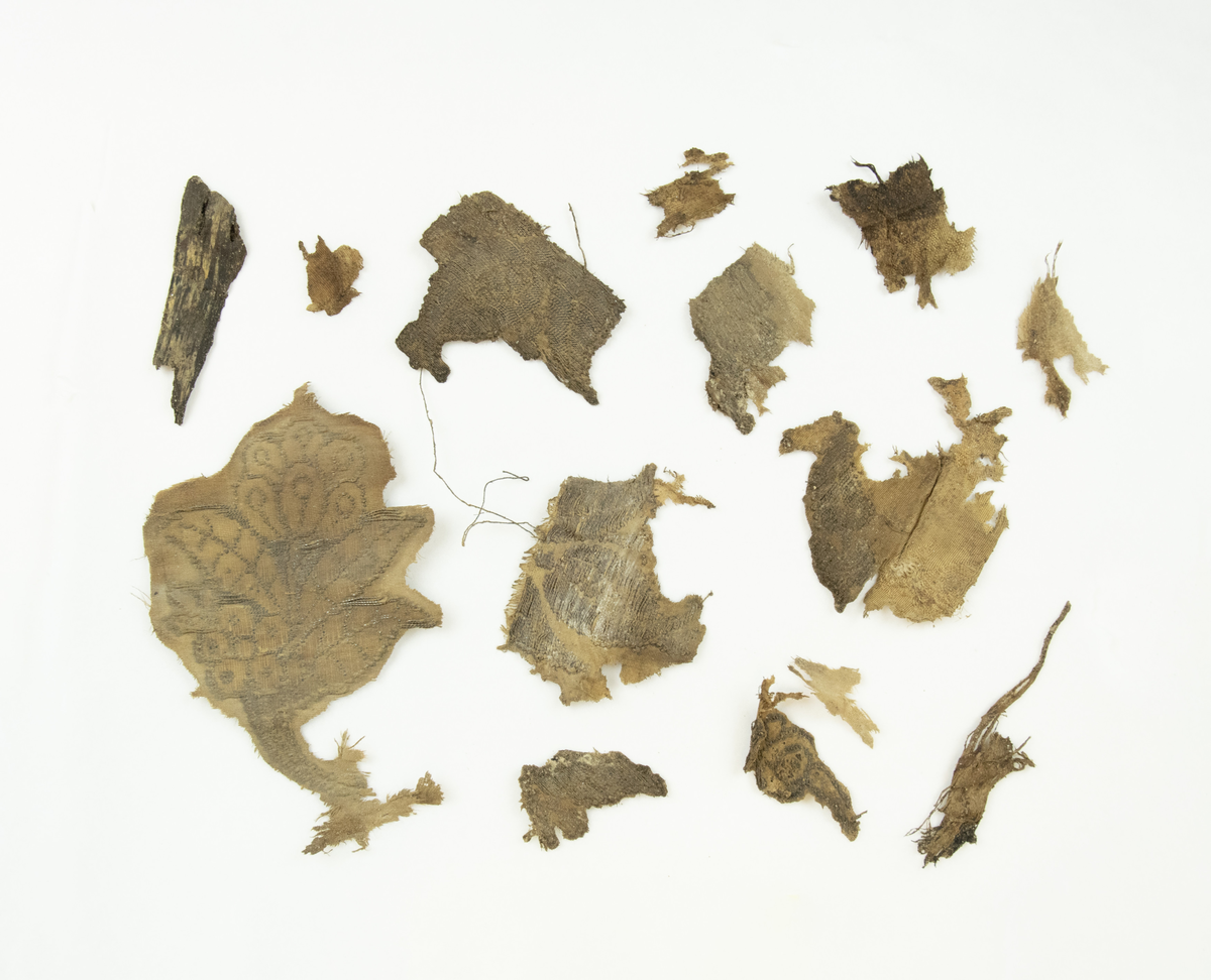 12 fragmenter av ulike størrelser og bevaringsgrad - det er bevart et nokså stort fragment med blomstermotiv, og flere av fragmentene er dekorerte. Fragmentene har antageligvis vært del av samme stykke stoff av ripsvev av silke- og/eller bomull og metalltråd. Samt fragmentene er det bevart to poser med vegetabilsk materiale funnet sammen med tekstilfragmentene. Materialet er foreløpig ukjent, men antatt å være planterester av humle, samt et trestykke som mulig kan være rest fra kisten, eller høvelflis som ikke var uvanlig å ha som underlag i kister på denne tiden. En pose med menneskehår er også bevart.