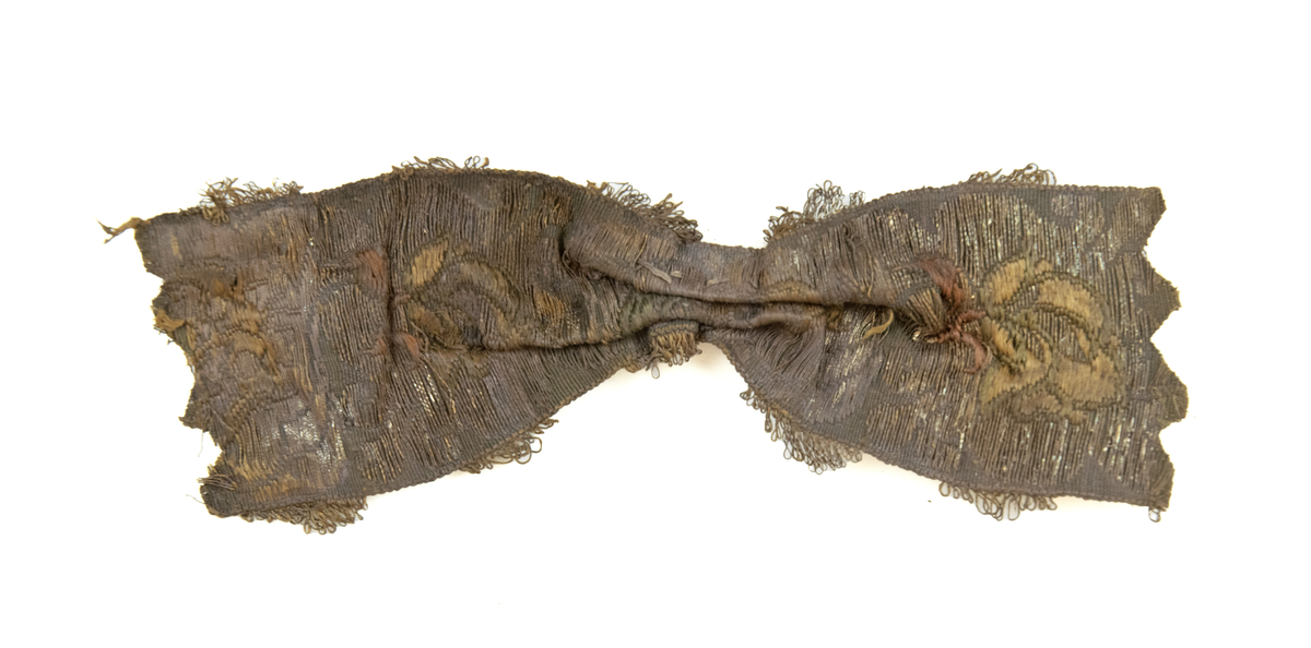 En sløyfe av brokade, antagelig av silkestoff og gull- og/eller tråd. Midten av sløyfen er presset og sydd sammen av tykk tråd (av bomull?). Den ene siden har grønt og rødaktig fargeskjær, de florale mønstrene er av metalltråd. Den andre siden har tydeligere fargesammensetninger i trådene - rød, gul og noe lilla. Løkker av metalltråd langs kantene. Taggete ender på sløyfa.

To lærfragmenter - den ene er avlang, og den andre tilspisset.