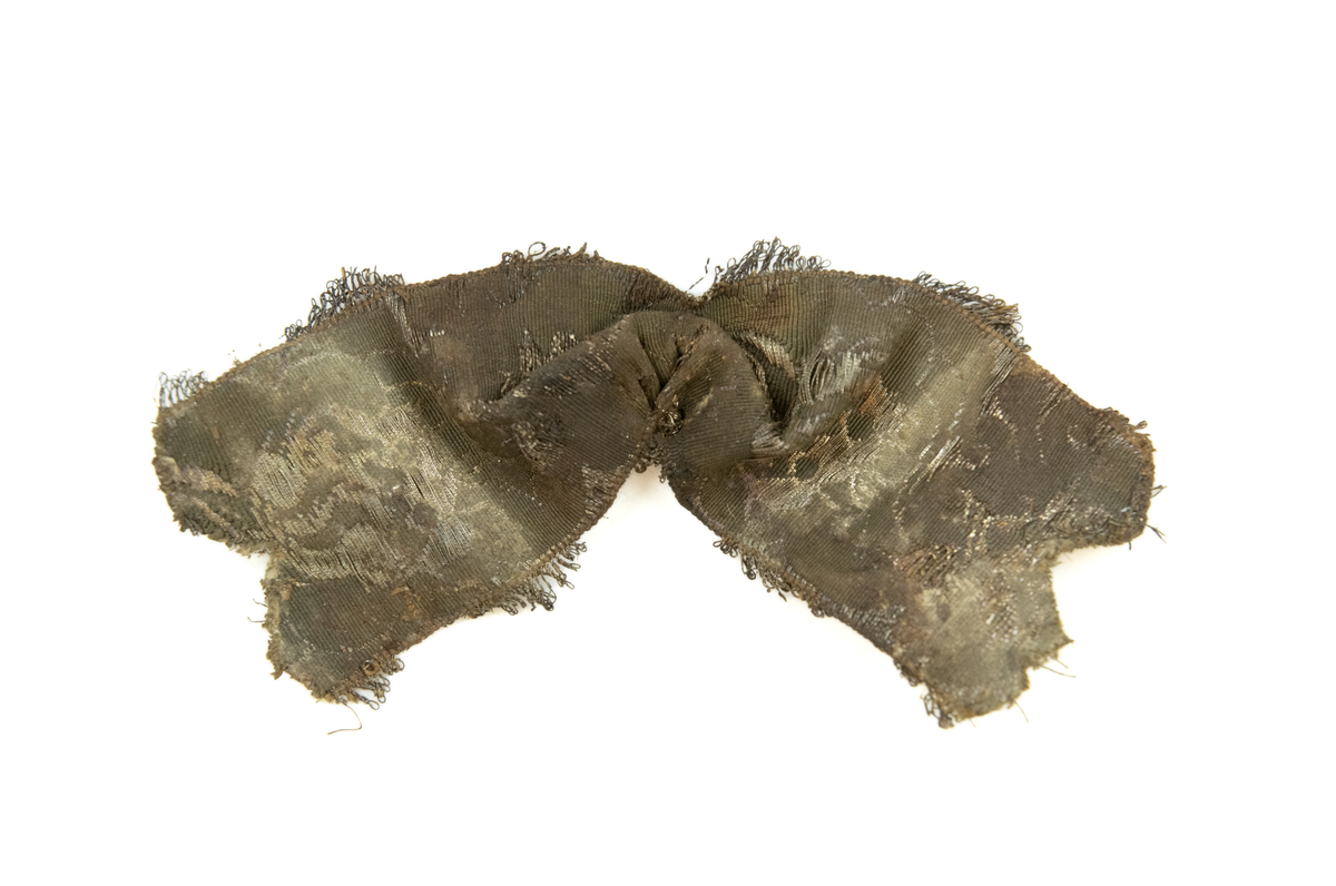 To deler bånd med tilhørende sløyfe. Vevd av silketråd, bomull(?), og metalltråder av antatt gull og sølv, med floralt mønster, typisk for rokokkoperioden. På den ene siden, antatt framsiden(?), er stoffet grønnfarget med florale motiver av metalltråd. På den andre siden, ses tydelige  fargede tråder av grønn, gul og rød silke/bomull.

Løkker av metalltråd, antatt sølv og/eller gull, langs begge langsidene av båndet. Gjennom hele båndet finner man rester av knappenåler av kobber som er korrodert. 

Sløyfen har tilnærmet lik mønstring og farger, og er antagelig formet av samme type bånd som hårbåndene.