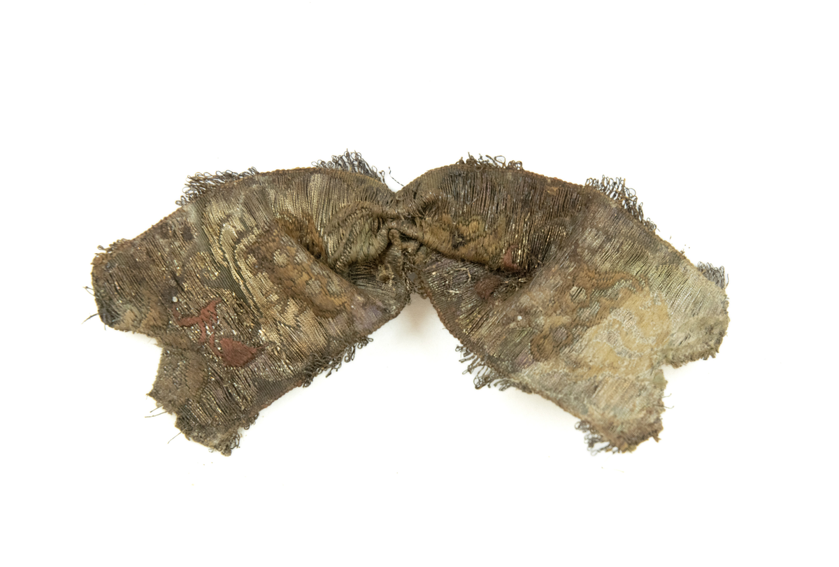 To deler bånd med tilhørende sløyfe. Vevd av silketråd, bomull(?), og metalltråder av antatt gull og sølv, med floralt mønster, typisk for rokokkoperioden. På den ene siden, antatt framsiden(?), er stoffet grønnfarget med florale motiver av metalltråd. På den andre siden, ses tydelige  fargede tråder av grønn, gul og rød silke/bomull.

Løkker av metalltråd, antatt sølv og/eller gull, langs begge langsidene av båndet. Gjennom hele båndet finner man rester av knappenåler av kobber som er korrodert. 

Sløyfen har tilnærmet lik mønstring og farger, og er antagelig formet av samme type bånd som hårbåndene.