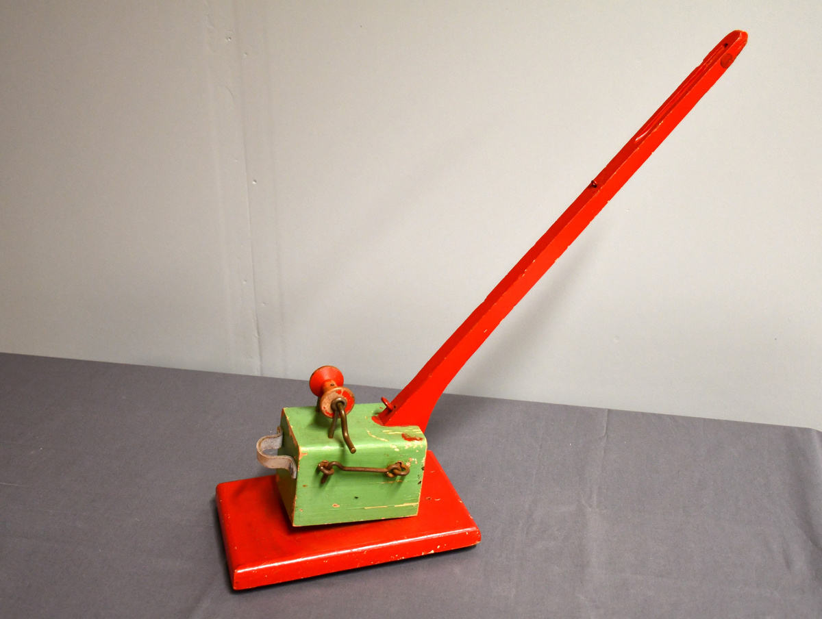 Rød og grønn høy kran i tre med bred base. Kan roteres 360 grader. Håndtakt i lær på bakparten, sveiv på toppen og hasp på høyre side.