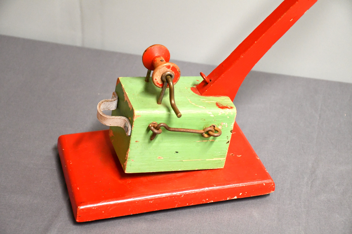 Rød og grønn høy kran i tre med bred base. Kan roteres 360 grader. Håndtakt i lær på bakparten, sveiv på toppen og hasp på høyre side.