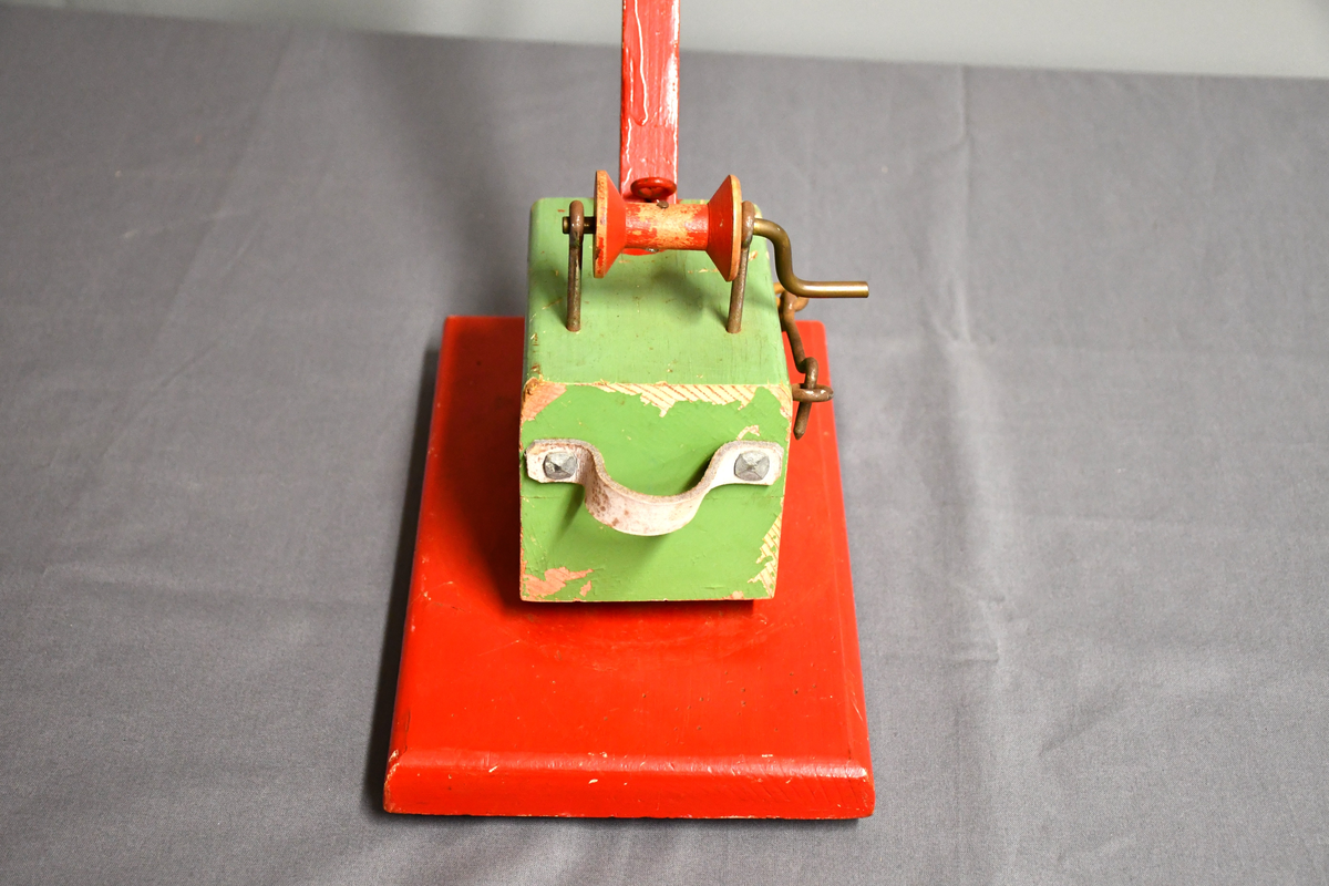 Rød og grønn høy kran i tre med bred base. Kan roteres 360 grader. Håndtakt i lær på bakparten, sveiv på toppen og hasp på høyre side.