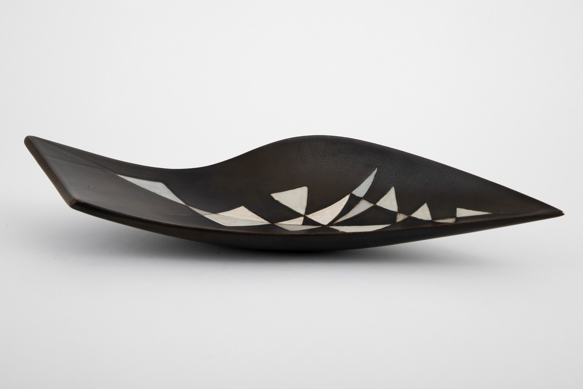 Fat i steingods. Uregelmessig, bøyd form, tilspisset i den ende enden og skrått avsluttet i den andre. Fatet er dekket med brunsvart glasur, innvendig dekorert med hvite og gråblå triangelfelt med innrissede konturer i et mosaikklignende mønster.