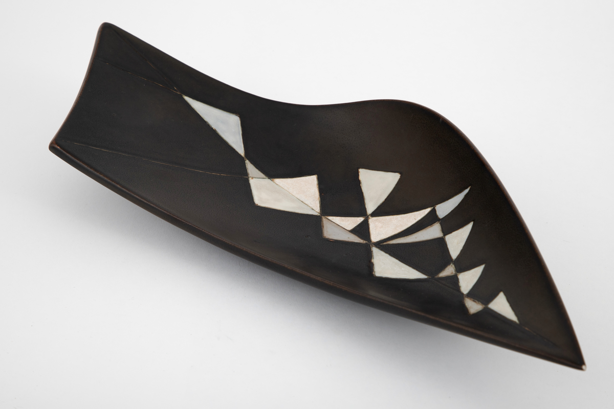 Fat i steingods. Uregelmessig, bøyd form, tilspisset i den ende enden og skrått avsluttet i den andre. Fatet er dekket med brunsvart glasur, innvendig dekorert med hvite og gråblå triangelfelt med innrissede konturer i et mosaikklignende mønster.