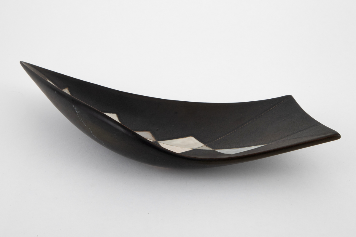 Fat i steingods. Uregelmessig, bøyd form, tilspisset i den ende enden og skrått avsluttet i den andre. Fatet er dekket med brunsvart glasur, innvendig dekorert med hvite og gråblå triangelfelt med innrissede konturer i et mosaikklignende mønster.