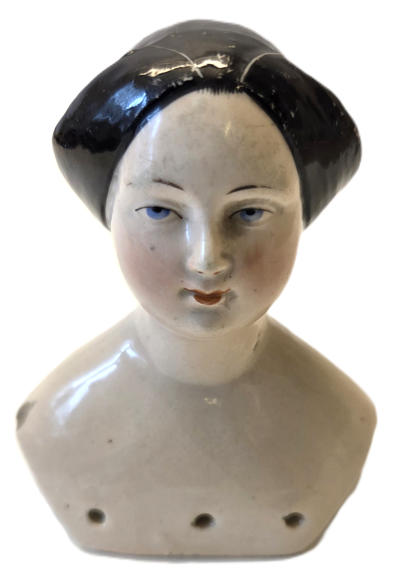 Porslin, flintgods? Glaserad rosa. Svart hår. Röda läppar. Blå ögon. Vit och blå etikett med "53". På insidan märkt "6v".
Gåva från Hedvig Ulfsparre.