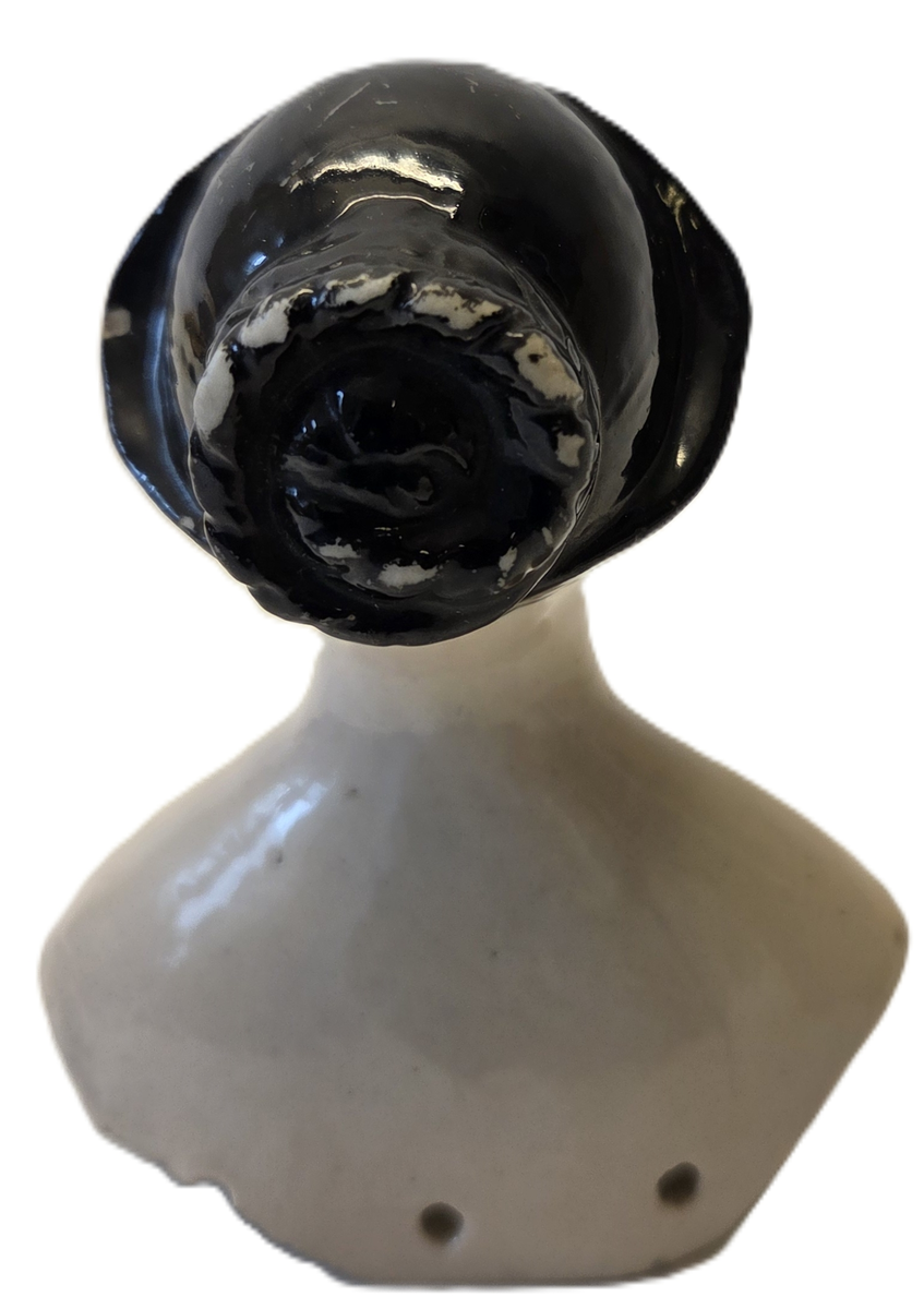 Porslin, flintgods? Glaserad rosa. Svart hår. Röda läppar. Blå ögon. Vit och blå etikett med "53". På insidan märkt "6v".
Gåva från Hedvig Ulfsparre.