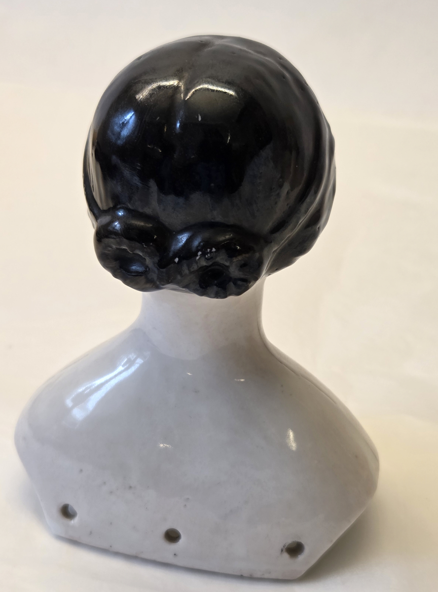 Porslin, vitglaserad. Rosa kinder. Svart hår. Blåa och svarta ögon. Röd mun. Vit och blå ettikett med "53".
Från Hedvig Ulfsparre.