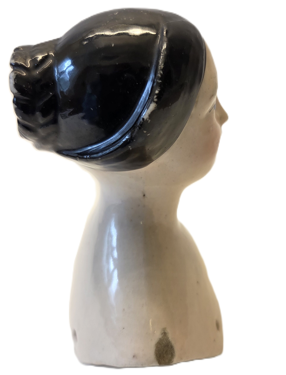 Porslin, flintgods? Glaserad rosa. Svart hår. Röda läppar. Blå ögon. Vit och blå etikett med "53". På insidan märkt "6v".
Gåva från Hedvig Ulfsparre.