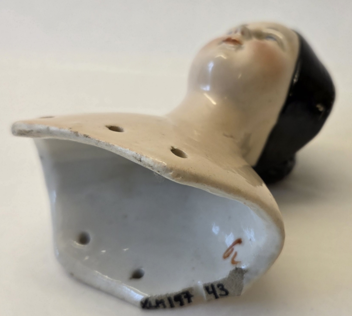 Porslin, flintgods? Glaserad rosa. Svart hår. Röda läppar. Blå ögon. Vit och blå etikett med "53". På insidan märkt "6v".
Gåva från Hedvig Ulfsparre.