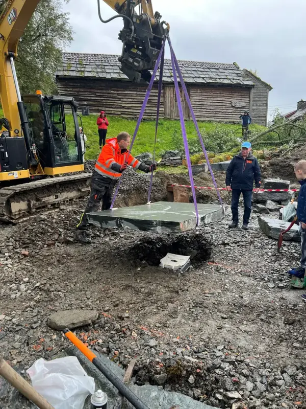 Grunnstein i huset legges ned ved gravemaskin. Menn i arbeidsklær står rundt. 