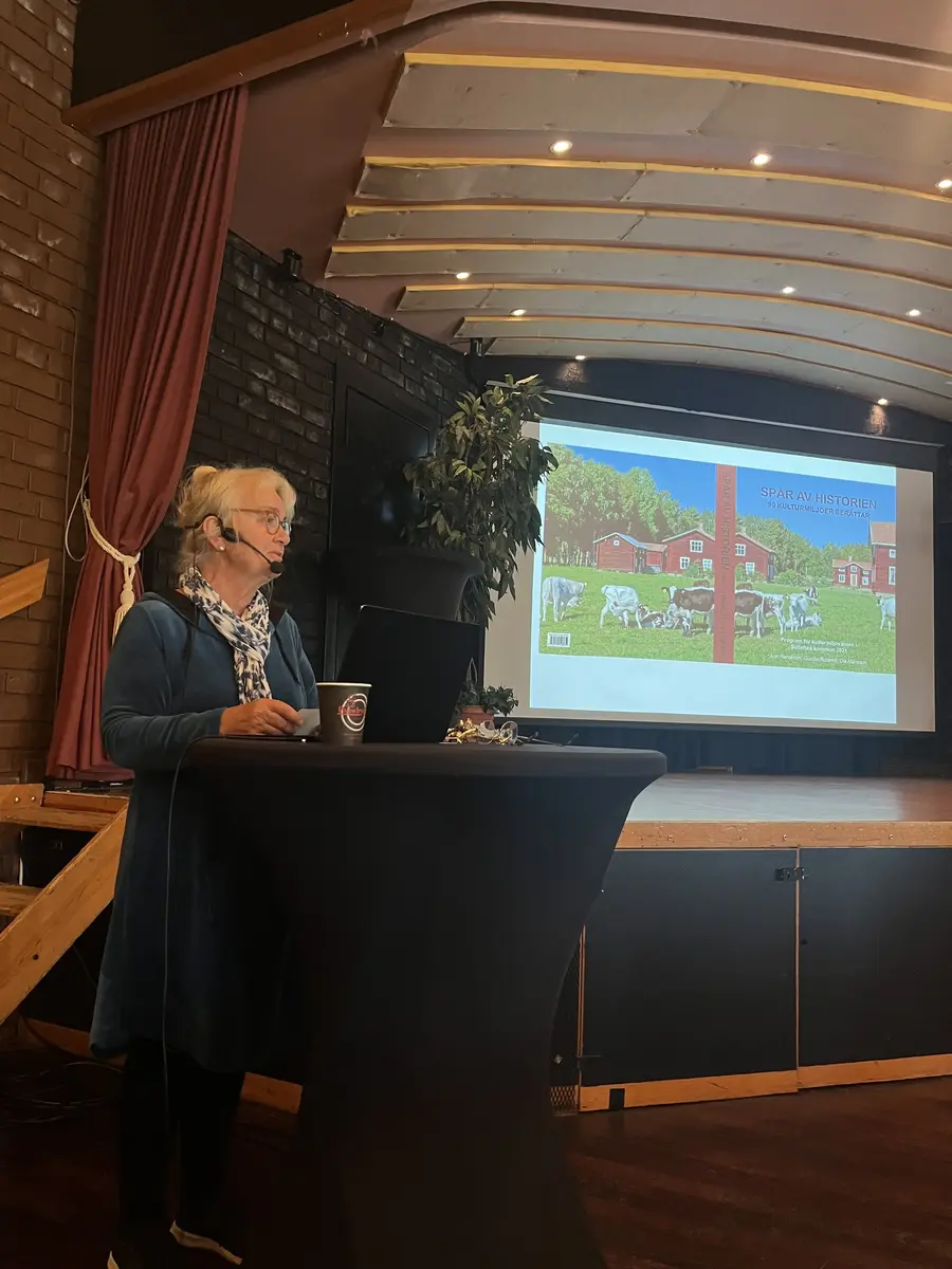 Kvinna som står vid podium med bildskärm i bakgrunden.