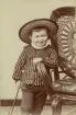 Foto ur John Herzbergs arkiv. 
Barnporträtt. Barnet poserar bredvid en korgstol. Barnet bär spetskrage och en skjorta med vertikala ränder, en stråhatt samt håller en stav i sin högra hand. Estemerad ålder på barnet: omkring 4-5 år.