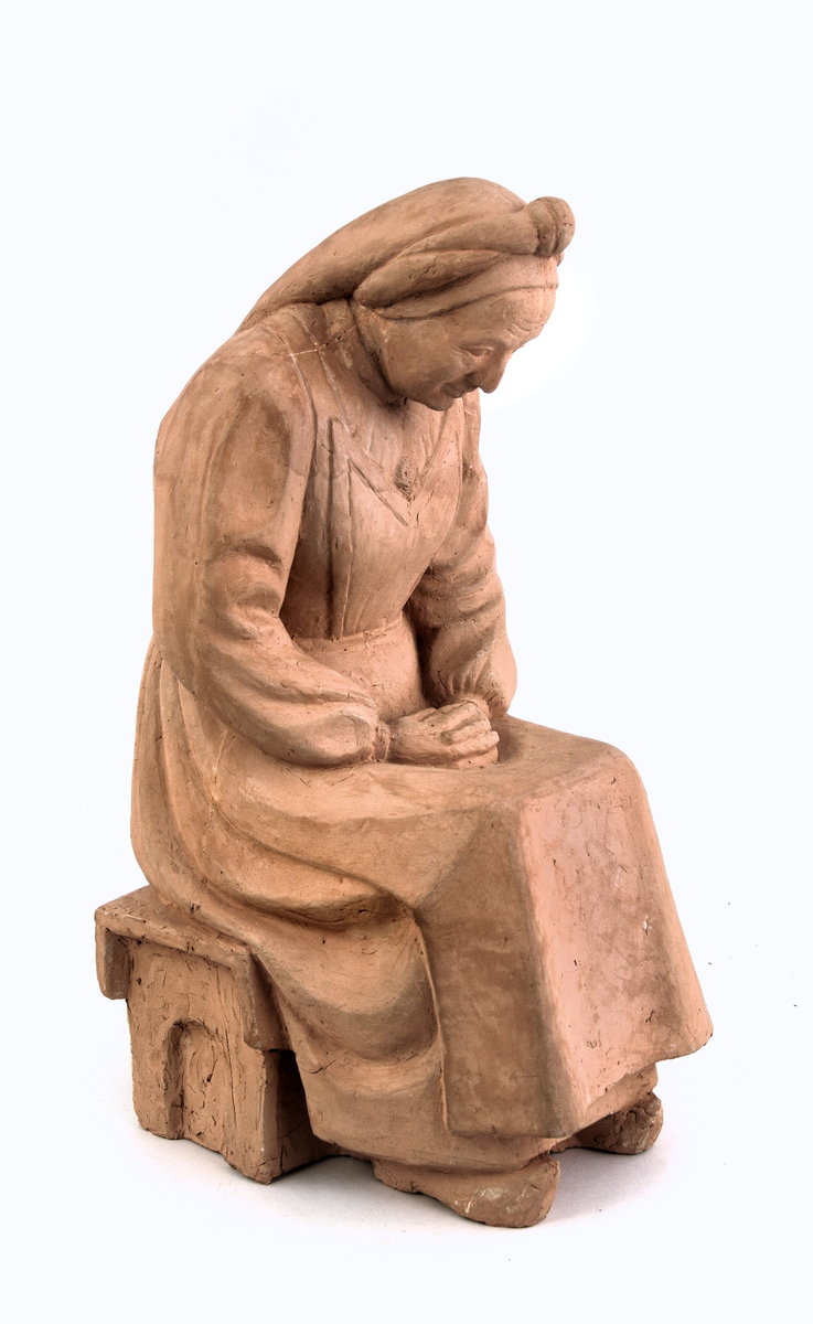 Gamal kvinne på skammel, kledd i stakk med bringeklut-trøye og tulleplagg, tresko på føtane, hendane læste i fanget. 