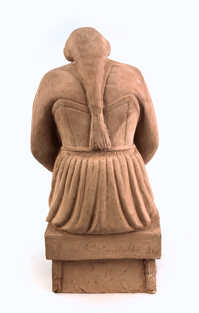 Gamal kvinne på skammel, kledd i stakk med bringeklut-trøye og tulleplagg, tresko på føtane, hendane læste i fanget. 