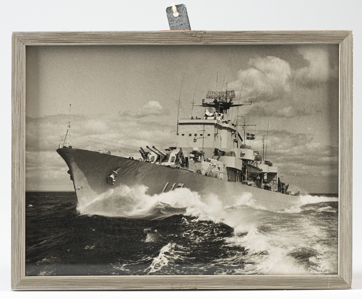 Svartvitt fotografi inom glas och ram visande jagaren Uppland under gång till havs, sedd från babord. Fotografiet är inramat i randad, gråmålad träram, B= 10mm. Baksidan klädd med kartong.