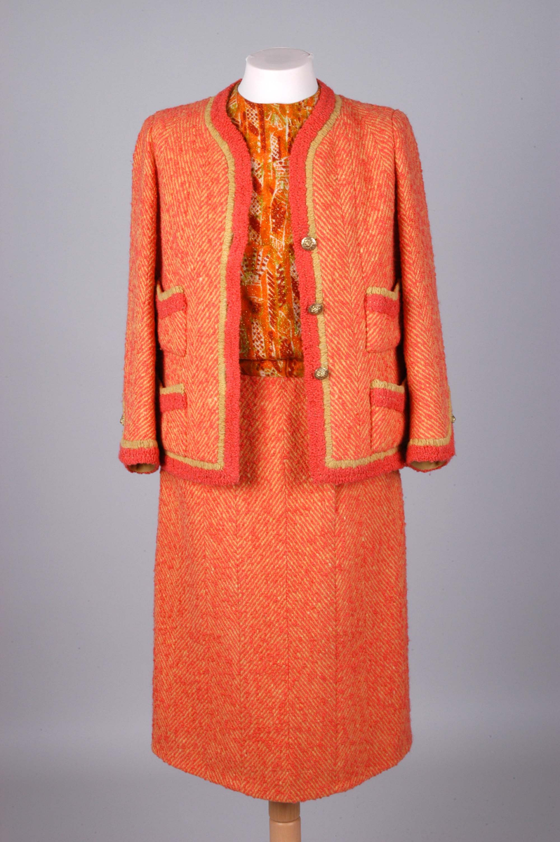 Todelt drakt bestående av kjole og jakke i korallfarget, fiskebensmønstret ull. Kjolen er konstruert slik at den ser ut som skjørt og bluse, og antrekket virker derfor tredelt. Det ermeløse kjolelivet har hel front og asymmetrisk lukking langs venstre skuldersøm og side. "Blusestoffet" er mønstret med gullfarget innslag. Skjørtet er fôret med silkestoff. Kjolelivet har svettelapper i silke. Jakken har pyntebånd i korall og gull, fire små lommer og gullfargede pynteknapper. Nederst på innsiden en påsydd gullfarget metallkjede. Fôret med korallfarget og sennepsgul silke.