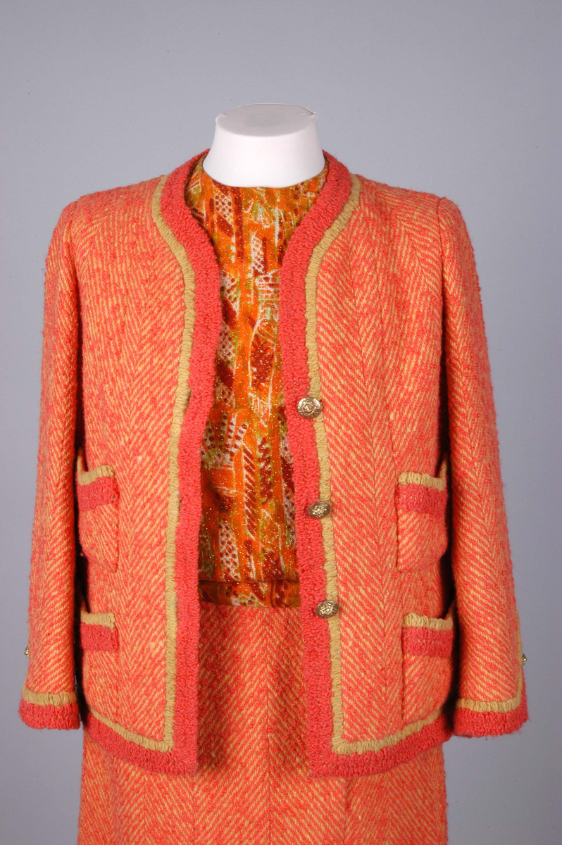 Todelt drakt bestående av kjole og jakke i korallfarget, fiskebensmønstret ull. Kjolen er konstruert slik at den ser ut som skjørt og bluse, og antrekket virker derfor tredelt. Det ermeløse kjolelivet har hel front og asymmetrisk lukking langs venstre skuldersøm og side. "Blusestoffet" er mønstret med gullfarget innslag. Skjørtet er fôret med silkestoff. Kjolelivet har svettelapper i silke. Jakken har pyntebånd i korall og gull, fire små lommer og gullfargede pynteknapper. Nederst på innsiden en påsydd gullfarget metallkjede. Fôret med korallfarget og sennepsgul silke.