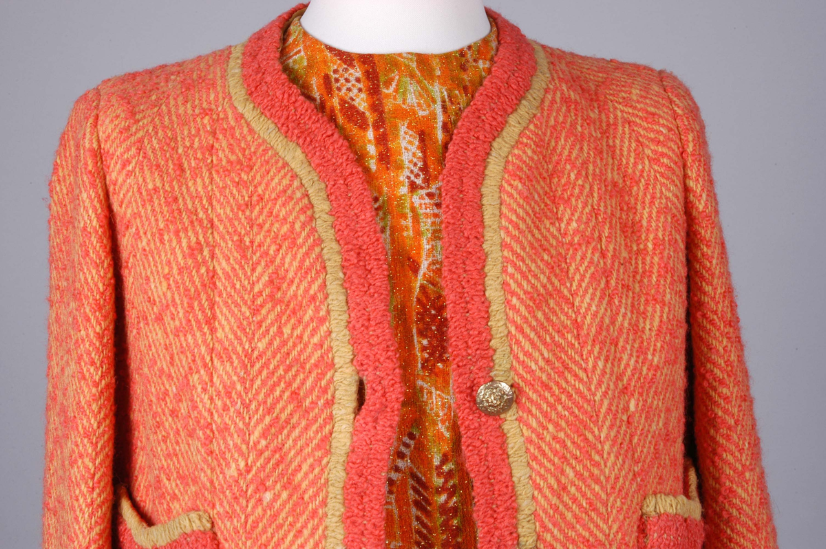 Todelt drakt bestående av kjole og jakke i korallfarget, fiskebensmønstret ull. Kjolen er konstruert slik at den ser ut som skjørt og bluse, og antrekket virker derfor tredelt. Det ermeløse kjolelivet har hel front og asymmetrisk lukking langs venstre skuldersøm og side. "Blusestoffet" er mønstret med gullfarget innslag. Skjørtet er fôret med silkestoff. Kjolelivet har svettelapper i silke. Jakken har pyntebånd i korall og gull, fire små lommer og gullfargede pynteknapper. Nederst på innsiden en påsydd gullfarget metallkjede. Fôret med korallfarget og sennepsgul silke.