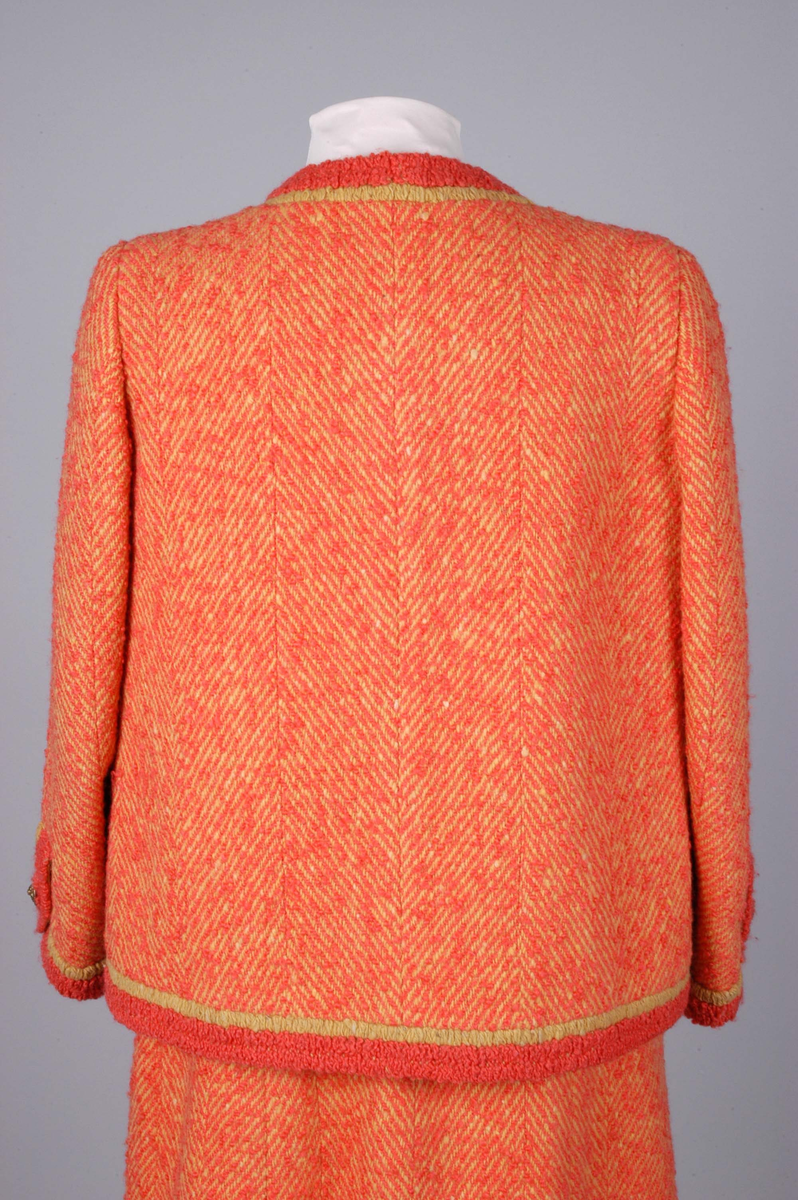 Todelt drakt bestående av kjole og jakke i korallfarget, fiskebensmønstret ull. Kjolen er konstruert slik at den ser ut som skjørt og bluse, og antrekket virker derfor tredelt. Det ermeløse kjolelivet har hel front og asymmetrisk lukking langs venstre skuldersøm og side. "Blusestoffet" er mønstret med gullfarget innslag. Skjørtet er fôret med silkestoff. Kjolelivet har svettelapper i silke. Jakken har pyntebånd i korall og gull, fire små lommer og gullfargede pynteknapper. Nederst på innsiden en påsydd gullfarget metallkjede. Fôret med korallfarget og sennepsgul silke.