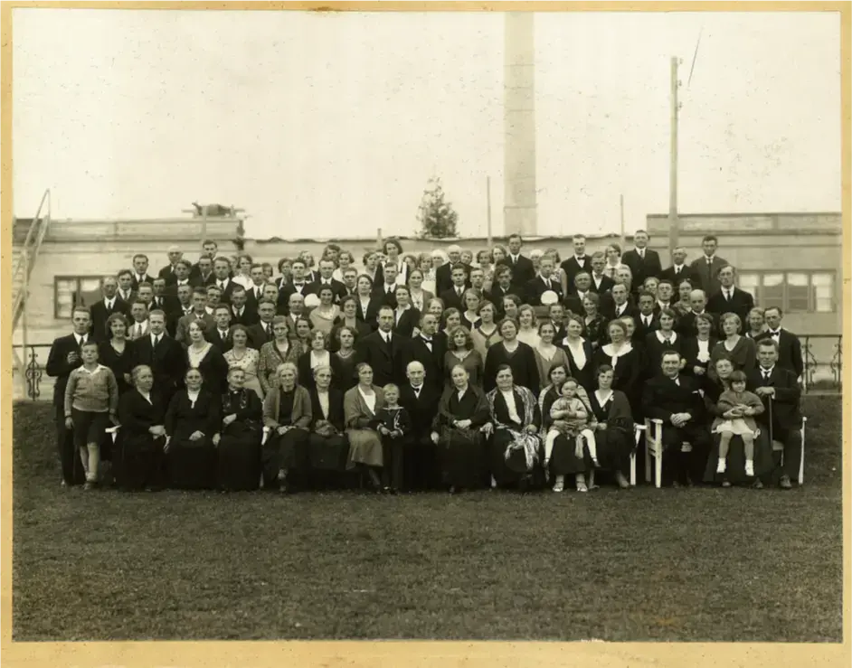 Bildet i sort-hvitt er et gruppebilde med mange mennesker stilt opp foran et fabrikkbygg med en høy pipe 