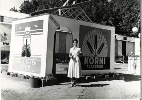 sort-hvitt-bilde av kvinne i kjole foran et enkelt bygg som ser ut som en flattbrødeske med dør i.