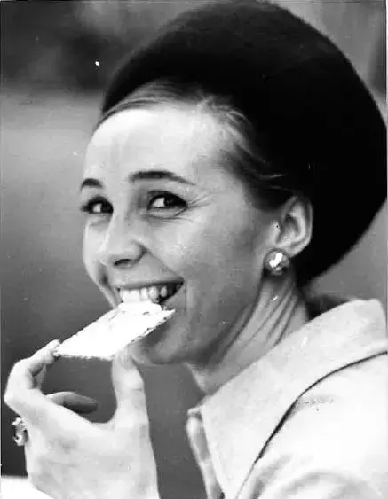 Sort-hvitt bilde av en pen kvinne med hatt og store øredobber. Hun holder et tynt knekkebrød opp mot munnen mens hun smiler og ser mot kameraet. 