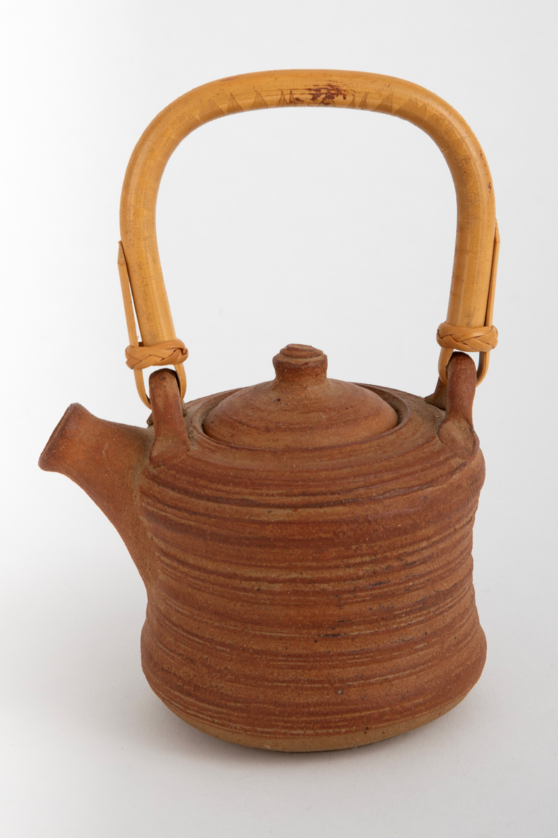 Liten brun tekanne med lokk og trehåndtak. Kraftige riller etter dreiingen. Noe konkav på midten av korpus.