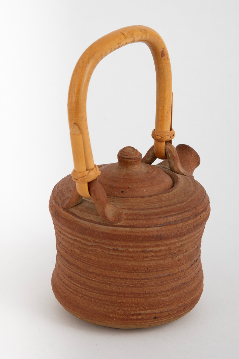 Liten brun tekanne med lokk og trehåndtak. Kraftige riller etter dreiingen. Noe konkav på midten av korpus.