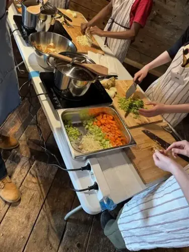 Her ser vi et bord med to kokeplater med store kjeler på, og barn som står rundt og kutter grønnsaker