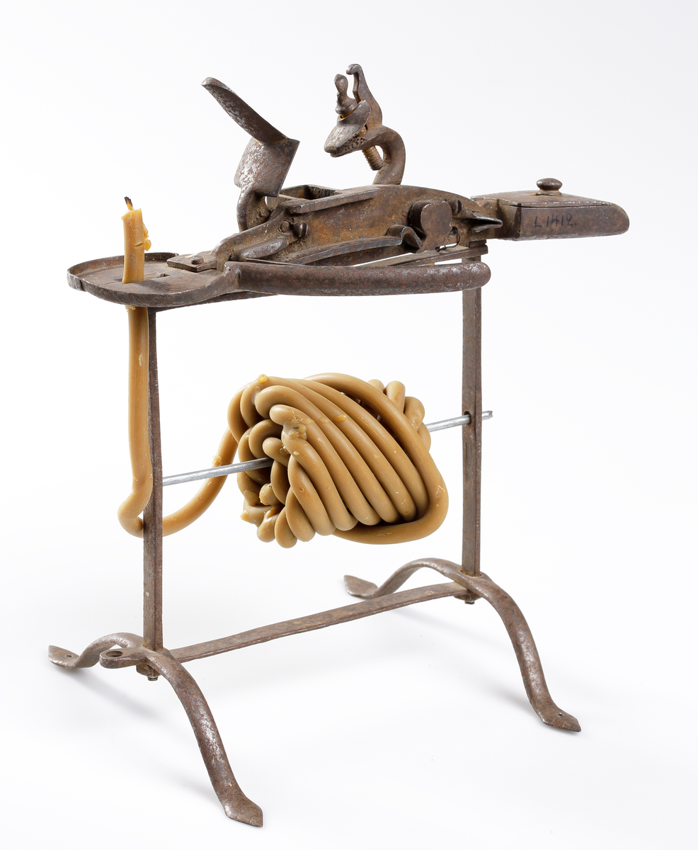 Vaxstapel eller järnställning för de förr brukliga rullarna av smala gula vaxljus, upptill med flintlås och antändningsapparat. Järncylindern varpå rullen fästades emellan två runda skivor (nu saknad),