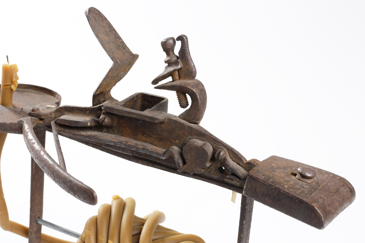 Vaxstapel eller järnställning för de förr brukliga rullarna av smala gula vaxljus, upptill med flintlås och antändningsapparat. Järncylindern varpå rullen fästades emellan två runda skivor (nu saknad),