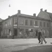 Korsningen Storgatan/Klostergatan (kv. Stormen) med bl a Lundbergs affär på hörnet, följt av Foto-Centralen och ingången till Vigfors matsalar.
Två herrar i kostym och hatt passerar.