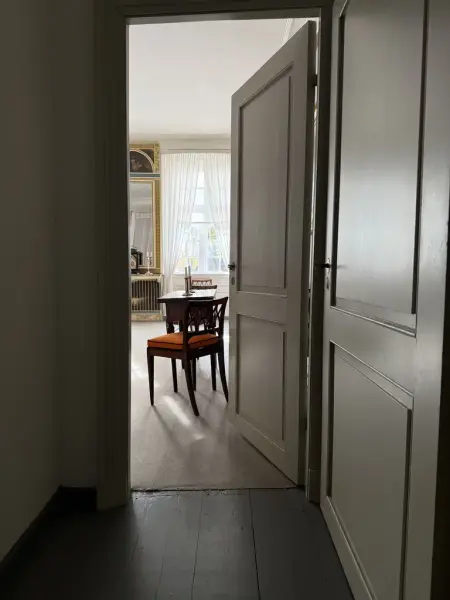 En åpen dør fra en gang inn til et lyst rom der man ser en stol og et speil på veggen. 