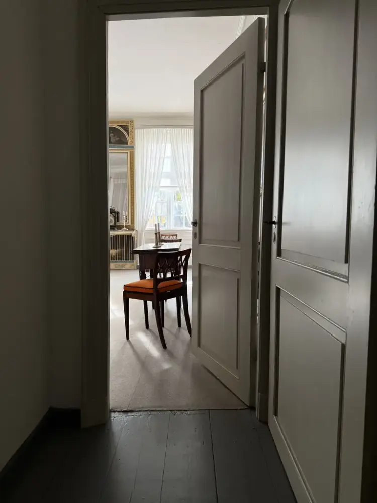 En åpen dør fra en gang inn til et lyst rom der man ser en stol og et speil på veggen. 