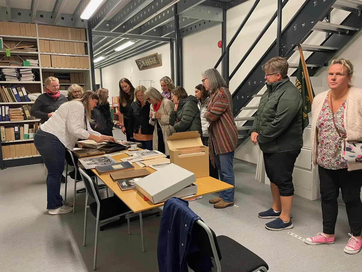 Bildet viser en gruppe kvinner som står ved et bord fylt av arkivmateriale (bilder og protokoller). De står inne i et arkivmagasin.