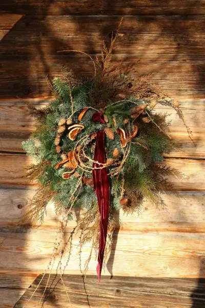 En grønn julekrans med kongler og rødt bånd henger på en brun tømmervegg. 