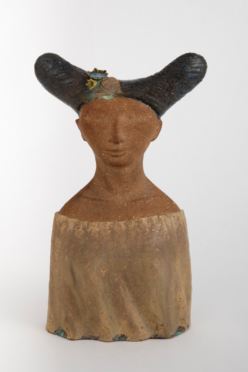 Skulptur i chamottekeramikk forestillende et kvinnehode en face. Ansikt og hals er uglasert, kjolen er dekket med gulbrun glasur. Håret, med en form som kan påminne om tre horn, er dekket med en dyp blå glasur og innrissede buede streker. I håret plastisk utformede blomster og blader i gult, blått og brunt.