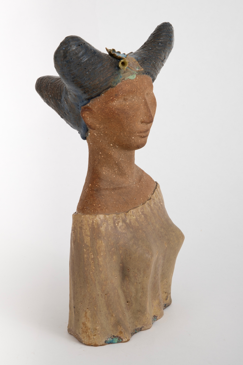 Skulptur i chamottekeramikk forestillende et kvinnehode en face. Ansikt og hals er uglasert, kjolen er dekket med gulbrun glasur. Håret, med en form som kan påminne om tre horn, er dekket med en dyp blå glasur og innrissede buede streker. I håret plastisk utformede blomster og blader i gult, blått og brunt.