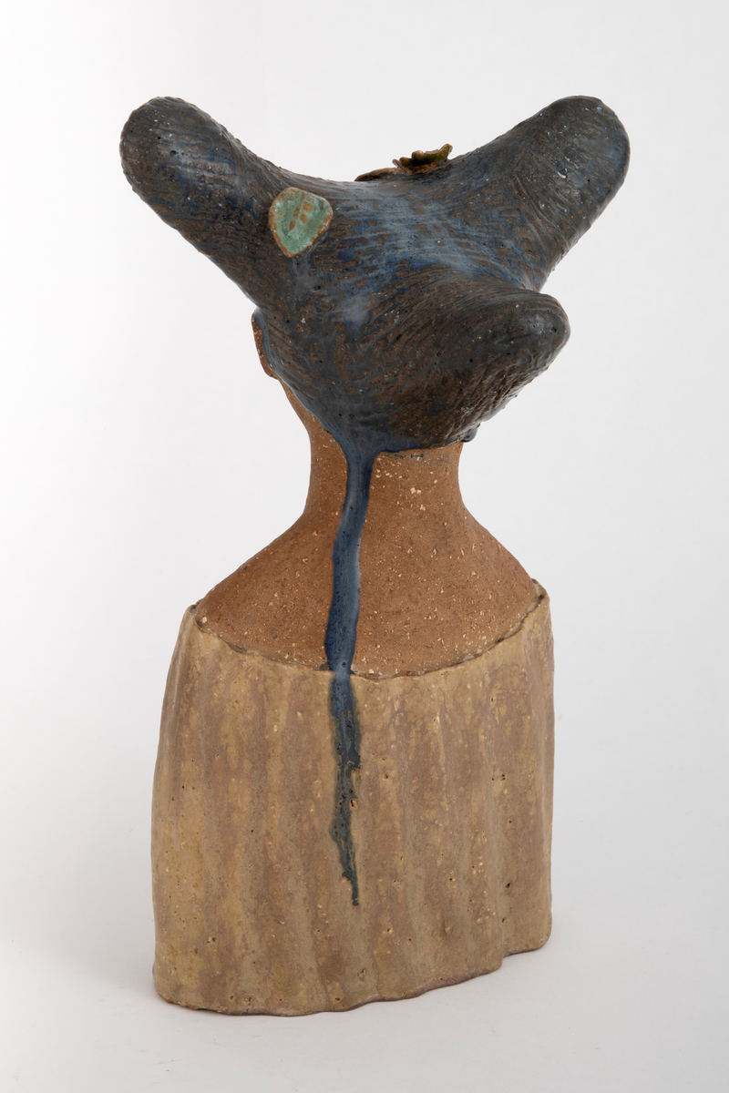 Skulptur i chamottekeramikk forestillende et kvinnehode en face. Ansikt og hals er uglasert, kjolen er dekket med gulbrun glasur. Håret, med en form som kan påminne om tre horn, er dekket med en dyp blå glasur og innrissede buede streker. I håret plastisk utformede blomster og blader i gult, blått og brunt.