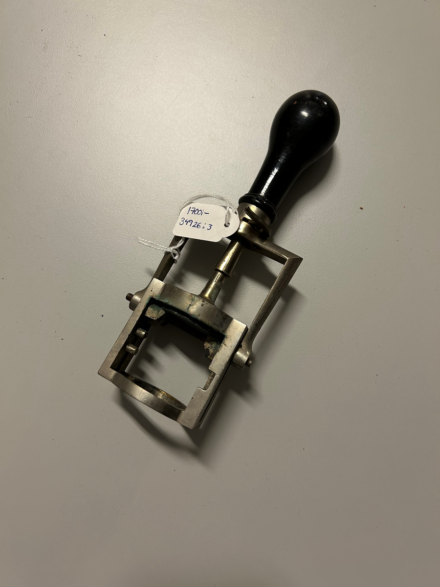 Ställning av metall igenom vilken en stämpel med handtag av svart trä är fäst. Stämpelns dyna av svart gummi med text utskuren i relief: "Karl Nyström. Kungl. hovfotograf Karlstad". Dynan doppas i stämpelfärg så att dynan då den trycks ned mot ett papper, återger texten. Dynan trycks ned genom att man trycker på handtaget och stämpeln förs nedåt med hjälp av att de skruvar som den är fastsatt med, löper i spår i ställningen.    Användes av brukaren för att märka de fotografier han framställde.