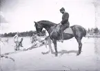Foto av en man i uniform som sitter på en häst vid en strand där ett gäng militärer tar sig ett bad. Text i album: 