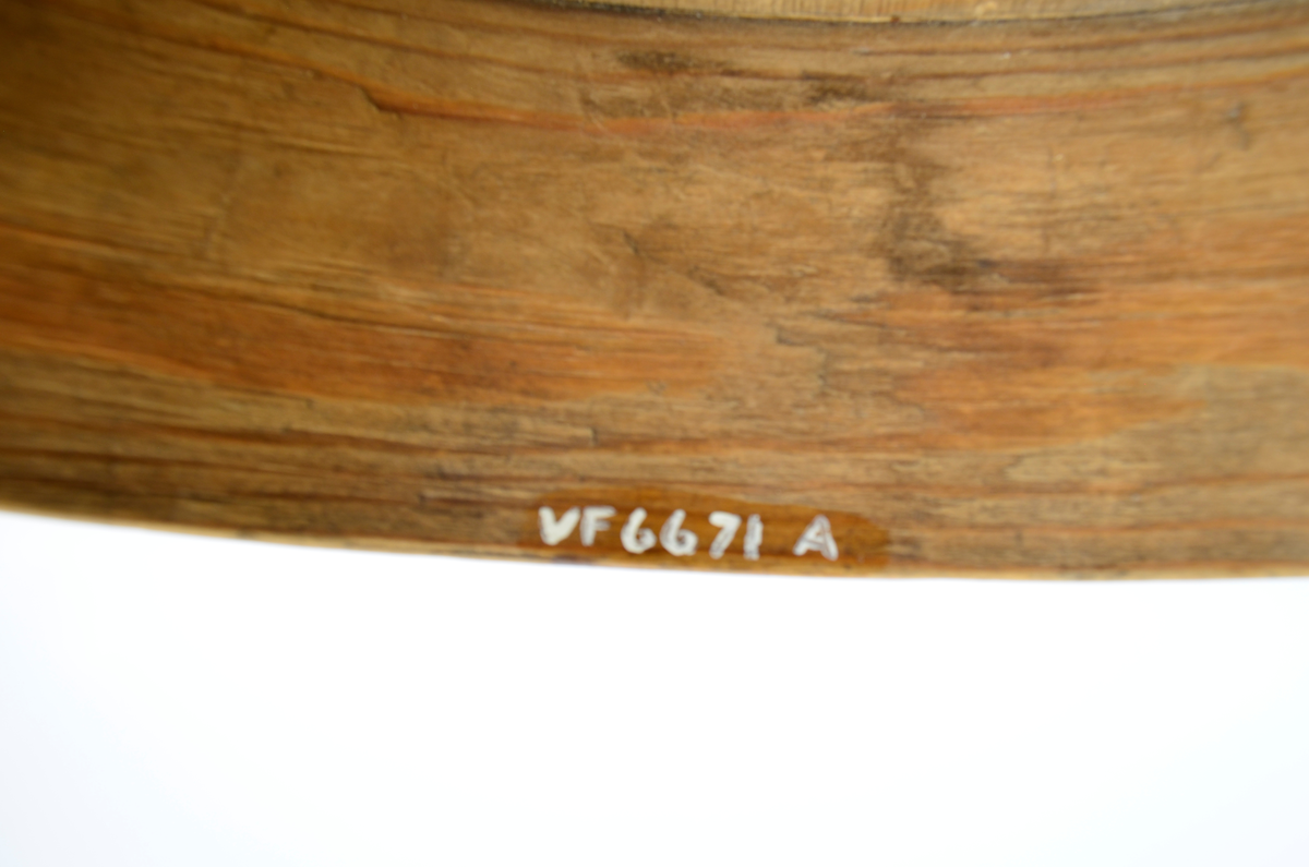 Gran. Sirkulær. Bunn og topp består av bord med spor langs kanten. Sidene, både i eske og lokk, utgjøres av bøyd spon, sydd sammen med tæger. Lokket er noe videre enn esken. Enkel sviornamentikk på innsiden av bunn og lokk, under bunnen og på sidene, rikere ornamentikk på lokkets overside. Sentralt sirkulært motiv, omgitt av fire kvadrater og fire rosetter. Buebord langs alle kanter.